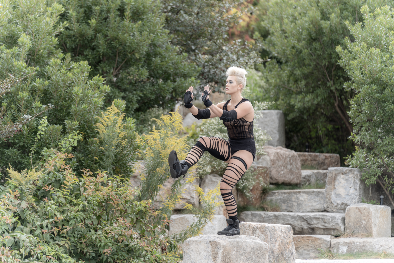 In ripped black tights and with a platinum blond punk hair, Robyn Cascio stands in profile on a slab of rocks.She has one leg bent and lifted. Both elbows are bent at right angles. 